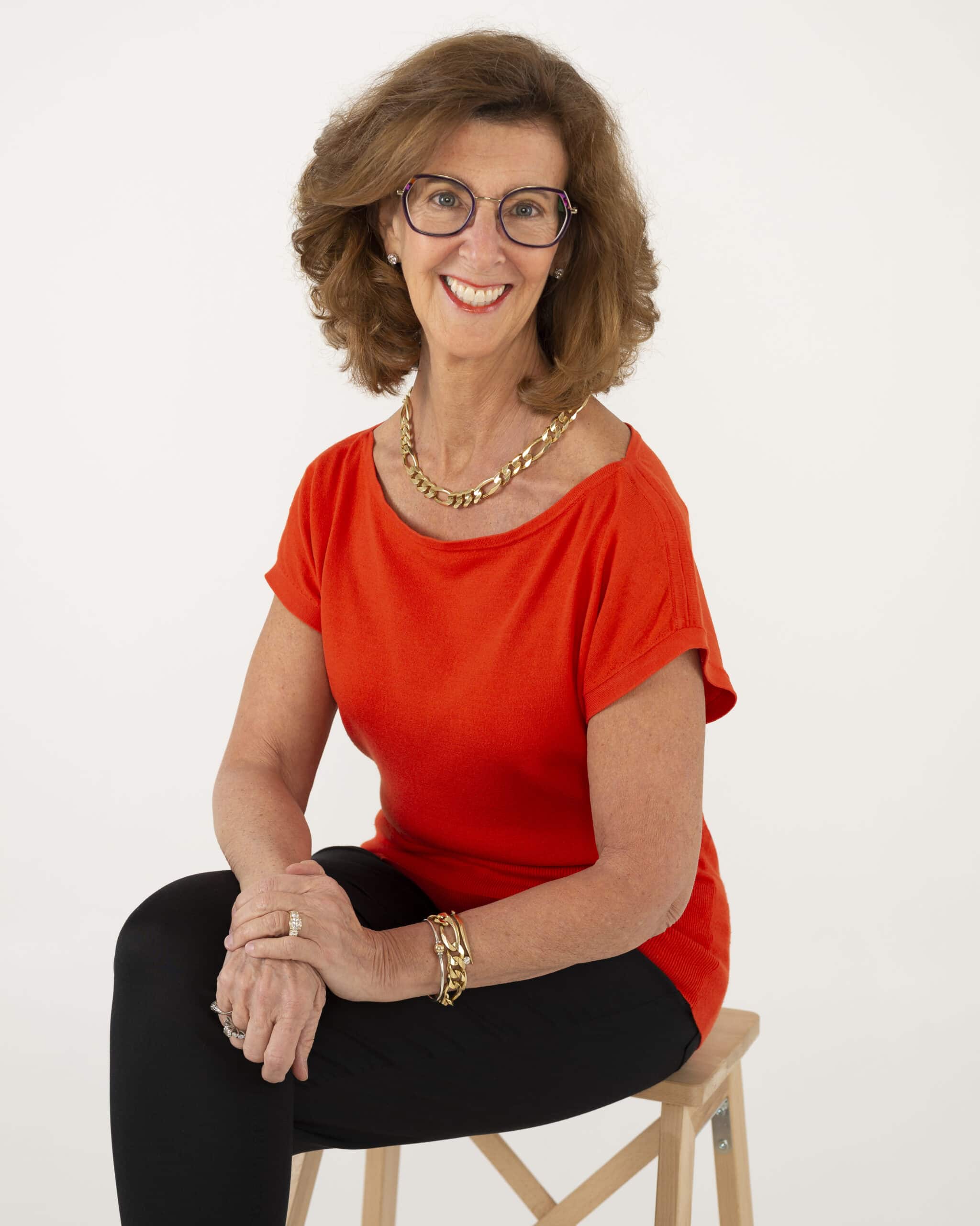 Image of Nancy Clairmont Carr sitting on a wooden stool with a red boat-neck shirt, gold necklace, and black slacks.